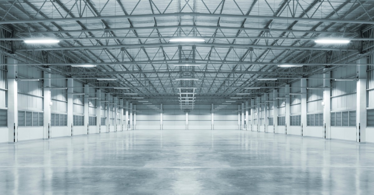 An empty industrial warehouse interior with polished concrete floors, metal wall panels, and rows of overhead lights.