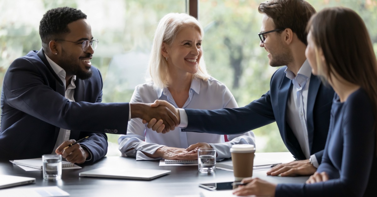 Four businesspeople sit around a conference table, with two shaking hands. Notebooks and beverages are on the table.