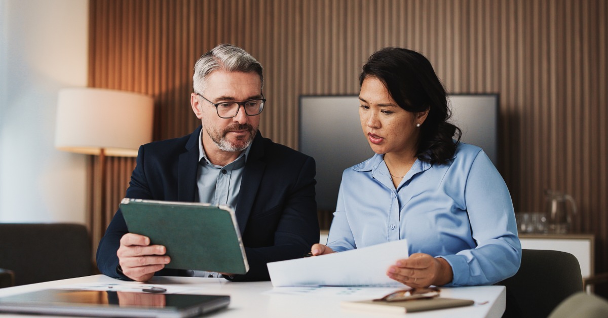 Two professionals sit at a table in an office, reviewing documents together. One holds a tablet, the other holds papers.