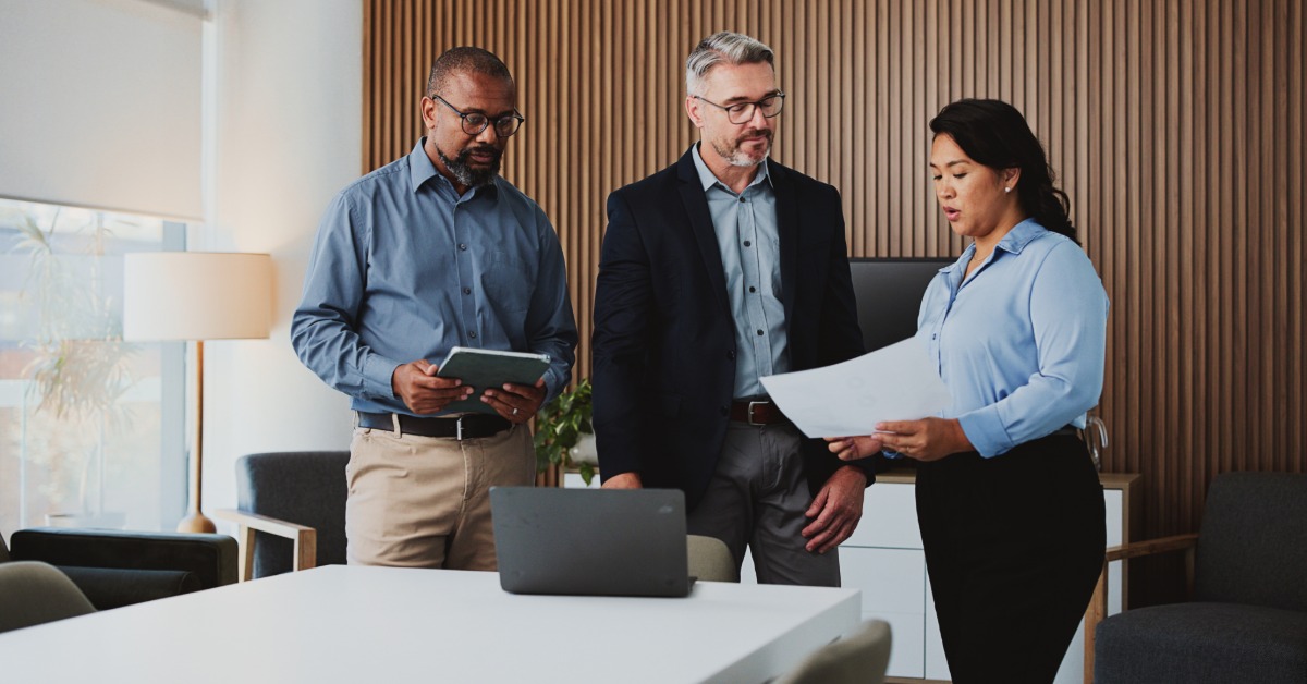 Three professionals review papers in an office with wood-paneled walls. An open laptop is on the table in front of them.