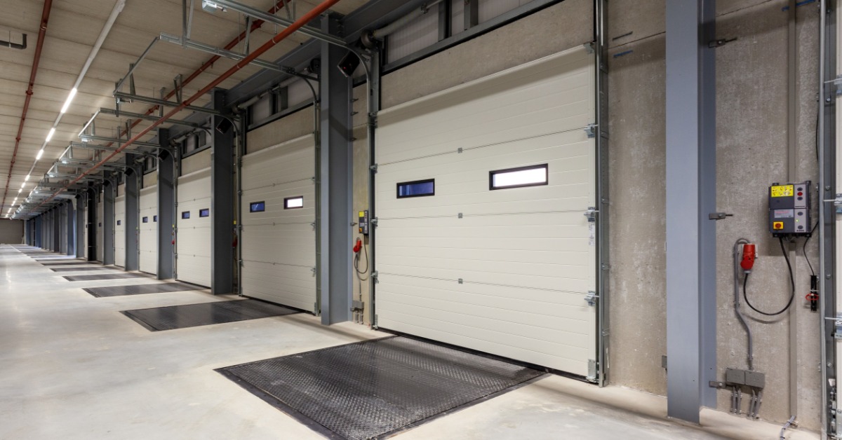 A row of white loading bay doors lines the inside wall of a warehouse. Black dock plates sit in front of each door.