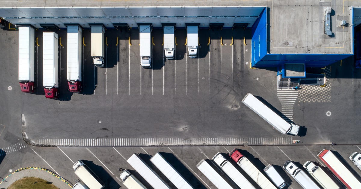 A warehouse loading area has multiple trailers backed into dock doors. More trucks and trailers are parked in the lot.