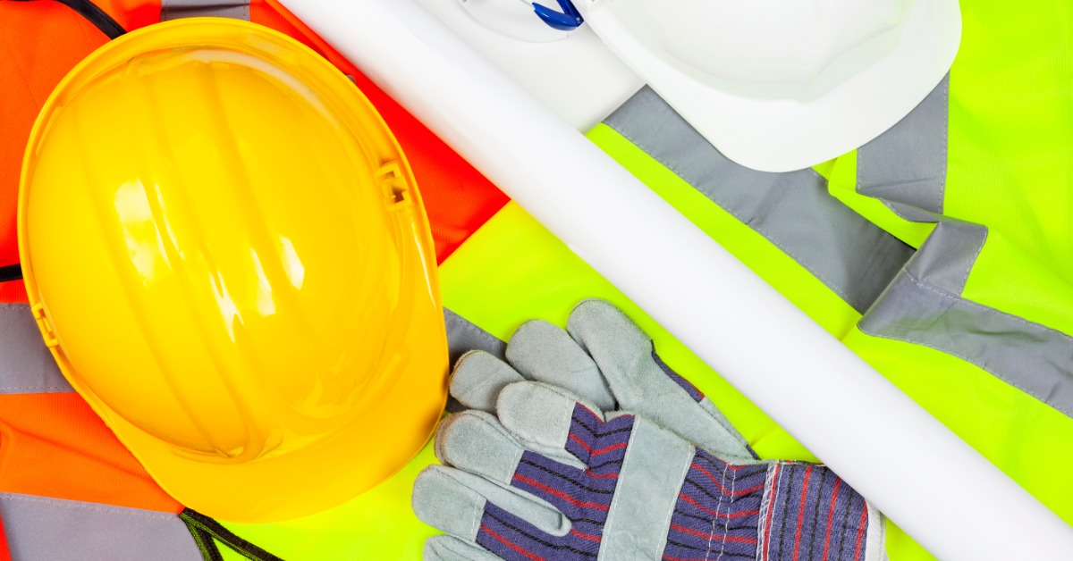 Hi-vis safety vests, a yellow hard hat, a white hard hat, work gloves, and safety glasses are arranged on a surface.
