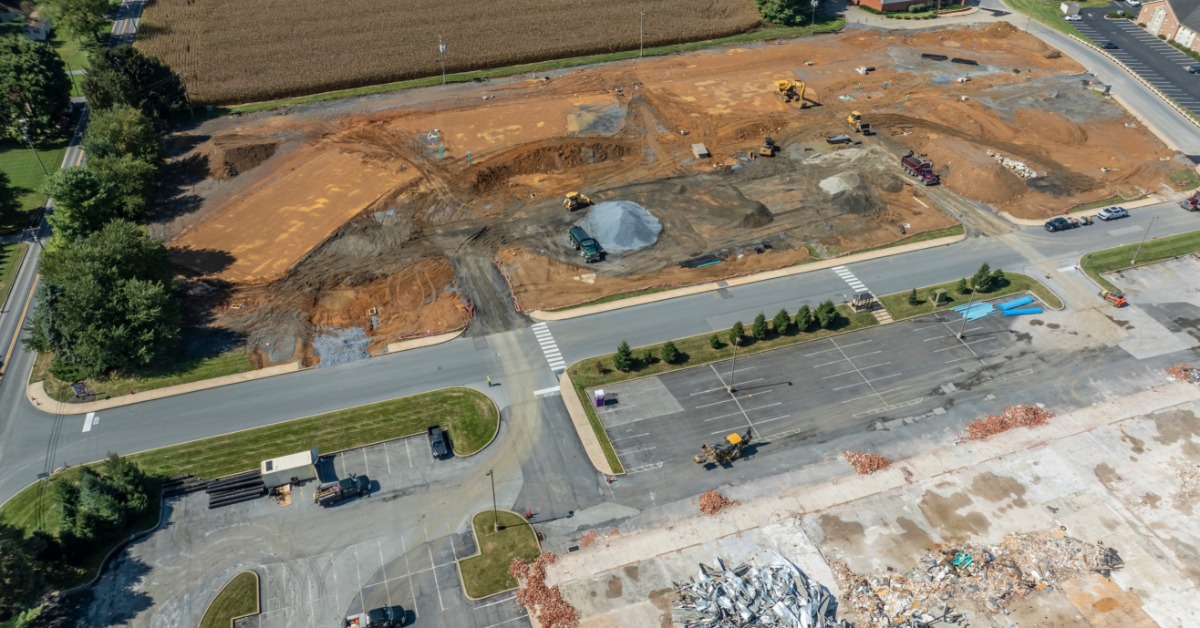 Heavy construction equipment works across a graded dirt parcel beside an existing roadway and parking areas.