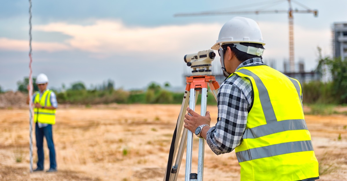A surveyor in a hard hat and reflective vest looks through a level on a tripod. Another worker holds a survey pole.