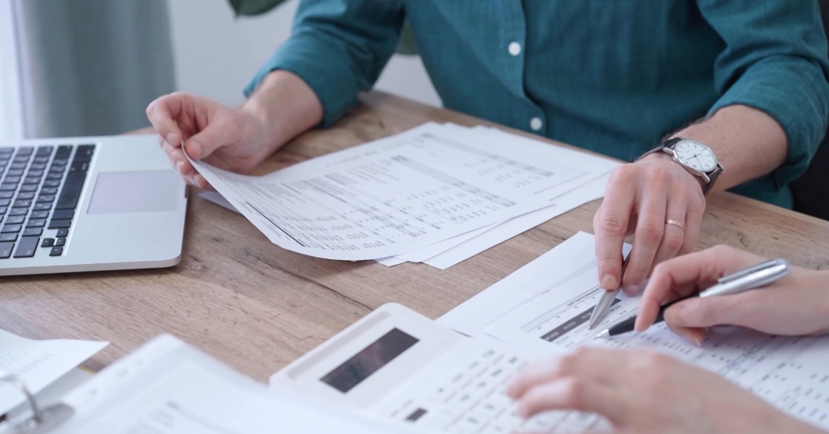 Two people holding pens review printed documents at a wooden table. A laptop and a calculator are also on the table.