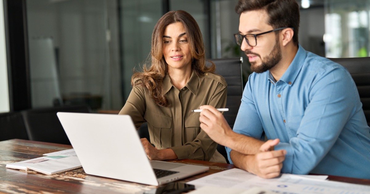 Two coworkers sit at a table in front of a laptop. The man holds a pen while the woman points at the computer screen.