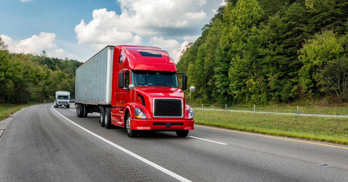 A red semi-truck pulls a silver trailer along a divided highway bordered by green trees. Another truck is behind it.
