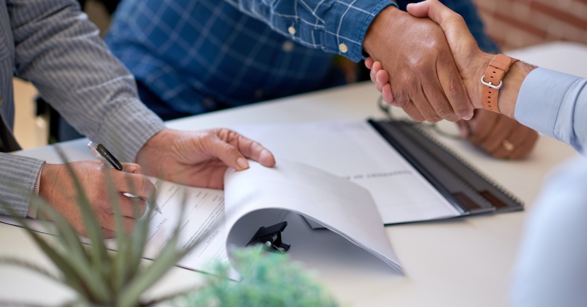 A close-up shows two people shaking hands above paperwork on a white table. A third person writes on a form.
