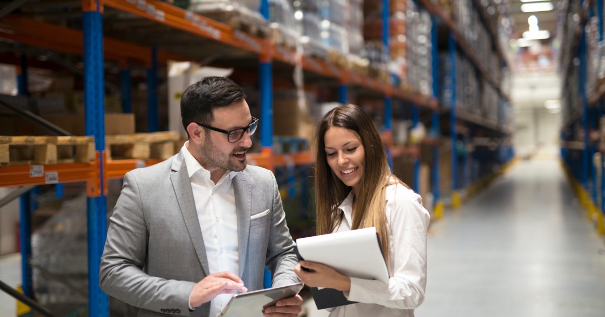 A man and woman in professional clothes look at paperwork and a tablet. They stand in front of racks in a warehouse.