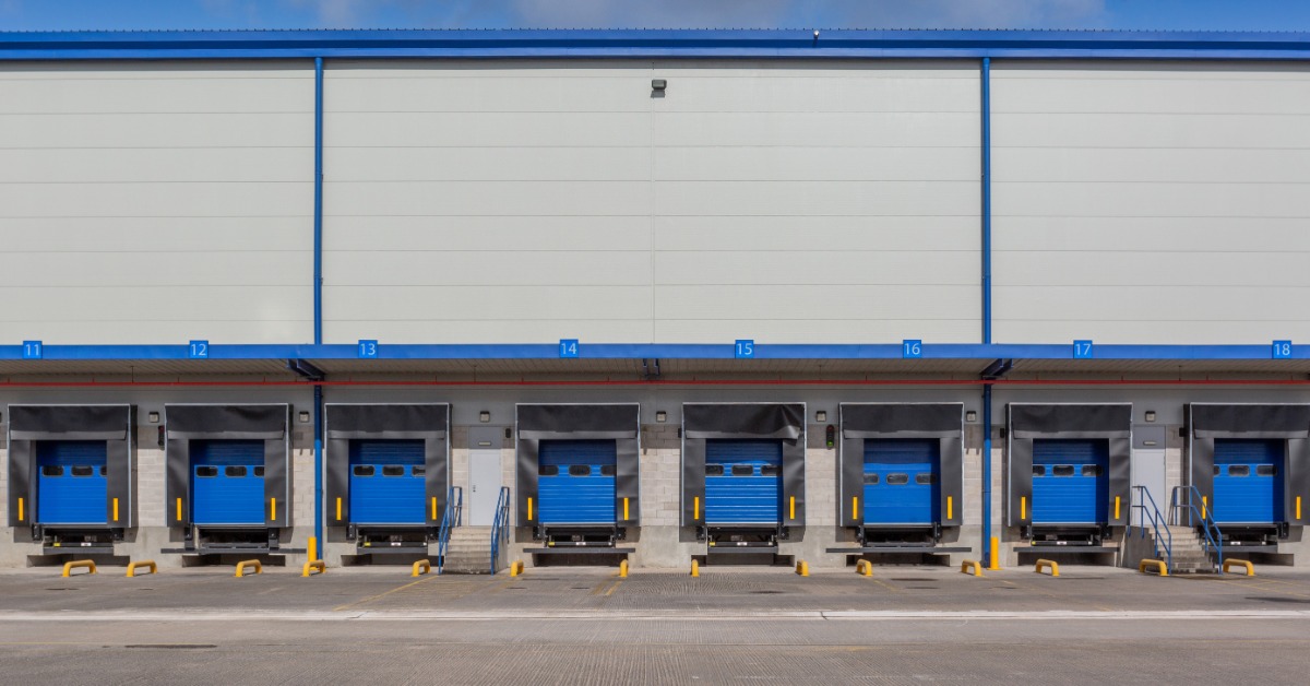 A row of blue loading dock doors lines the exterior of a warehouse. Yellow safety bollards are in front of the bays.