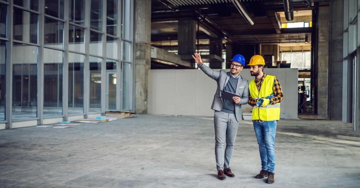 Two people wearing hard hats are in an unfinished industrial building. One holds a tablet and gestures toward the room.