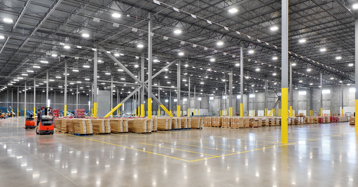 Palletized boxes sit in a large warehouse with high metal ceilings, bright overhead lights, and rows of support columns.