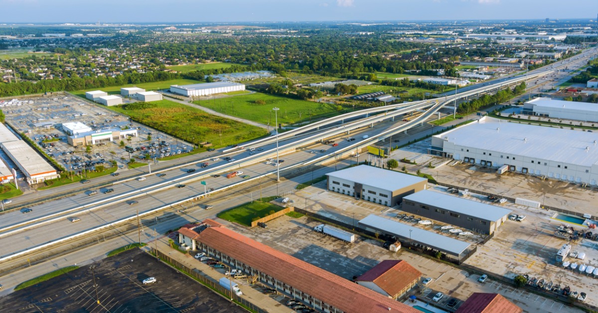 An aerial view shows several industrial buildings beside a large highway interchange with multiple lanes of traffic.