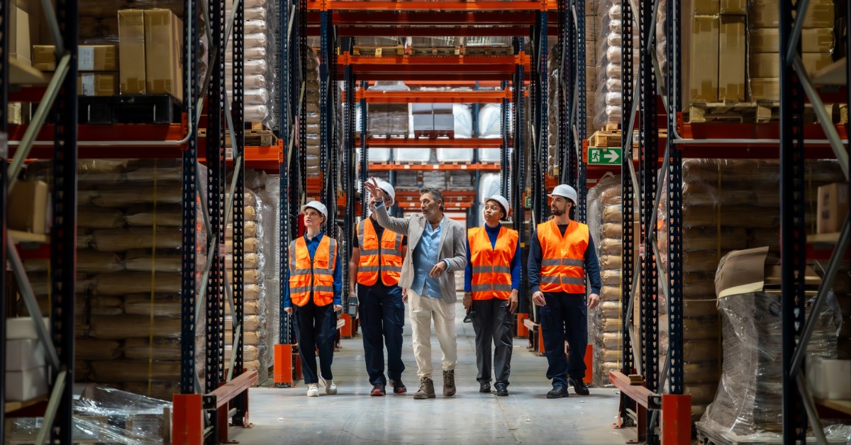 Four people in orange safety vests and a man in a suit walk together down a warehouse aisle between tall storage racks.