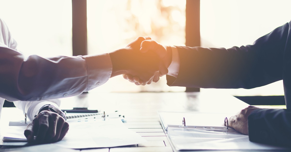 Two people shake hands across a table covered with documents. Bright sunlight shines through the windows behind them.