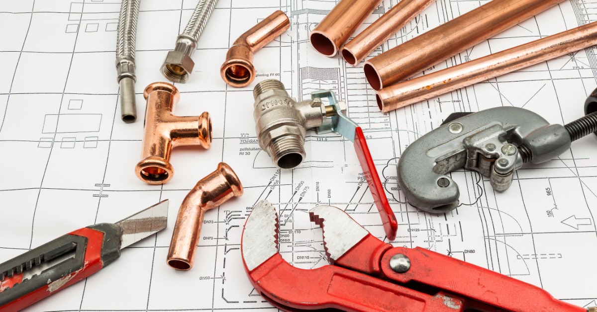 Plumbing materials and tools rest on top of architectural plans, including copper tubing, valves, and metal hoses.