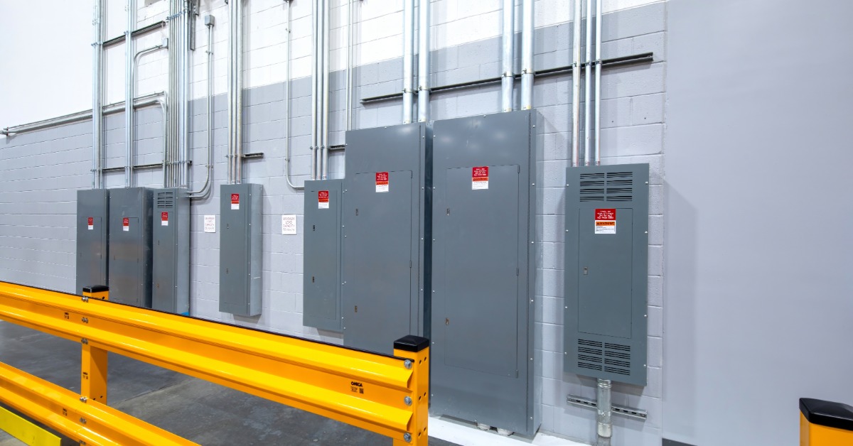 Gray electrical panels and metal conduit are mounted along a light gray industrial wall behind yellow guardrails.