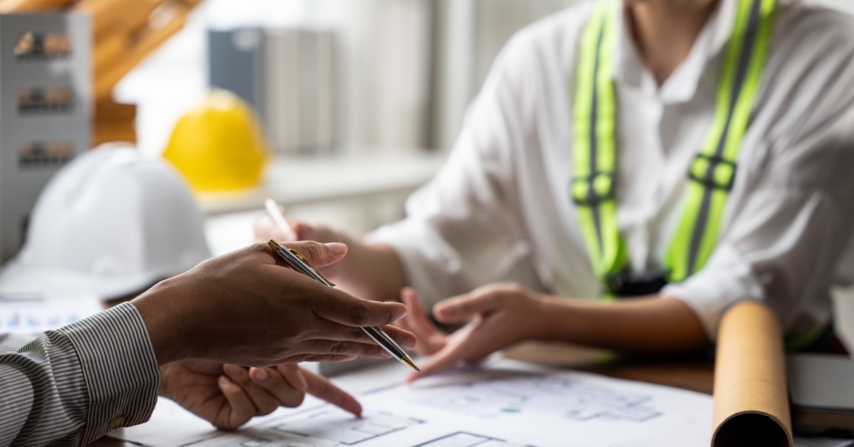 Two people sit at a table with floor plans. One wears a reflective safety vest while the other points to the plans.
