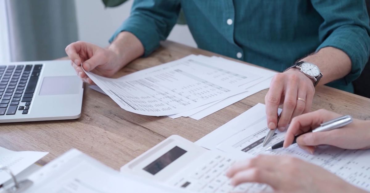 Two people review paperwork together at a desk with a laptop and a calculator. They point to figures on a page.
