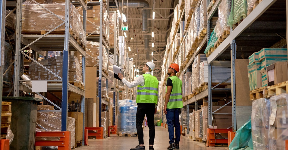 Two warehouse workers wearing high-visibility vests and hard hats stand in an aisle between tall storage racks.