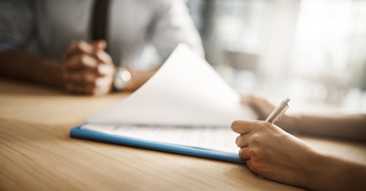 Two people sit across from each other at a table with documents. One lifts some of the paper and signs a page.