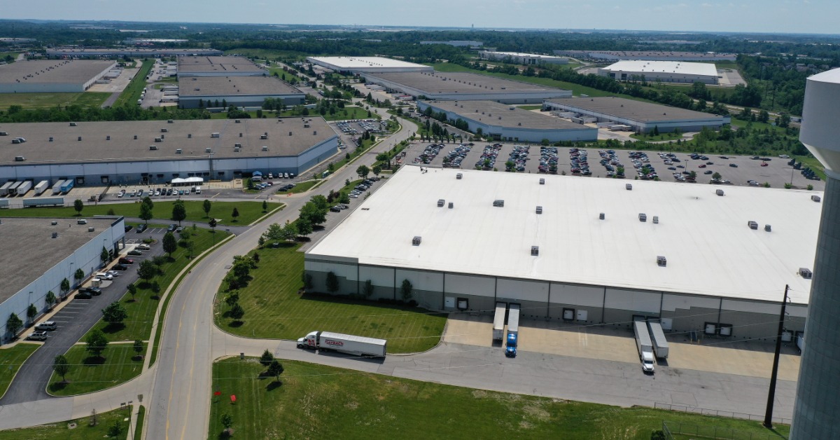 An aerial view of several warehouses with loading docks, parking lots, and access roads. A truck leaves a loading area.