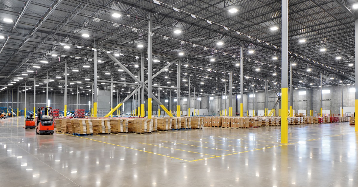 A spacious warehouse has exposed steel beams and bright overhead lights. Palletized goods sit near parked forklifts.